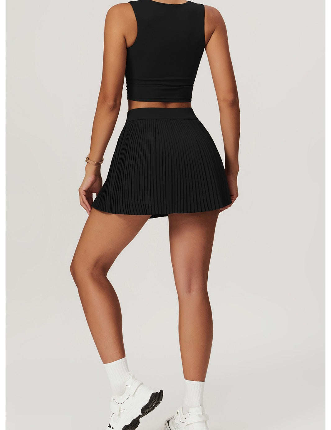 Person wearing a black sleeveless top and pleated skort with white sneakers on a light gray background